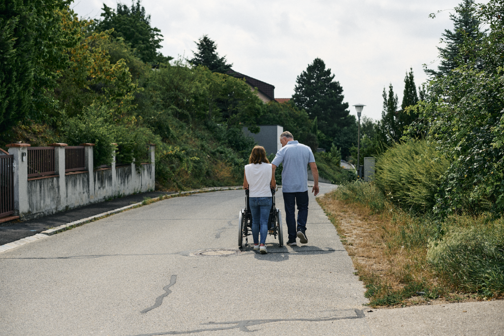Ein älterer Mann und eine Frau schieben gemeinsam einen Rollstuhl auf einer Straße umgeben von Bäumen und Sträuchern.