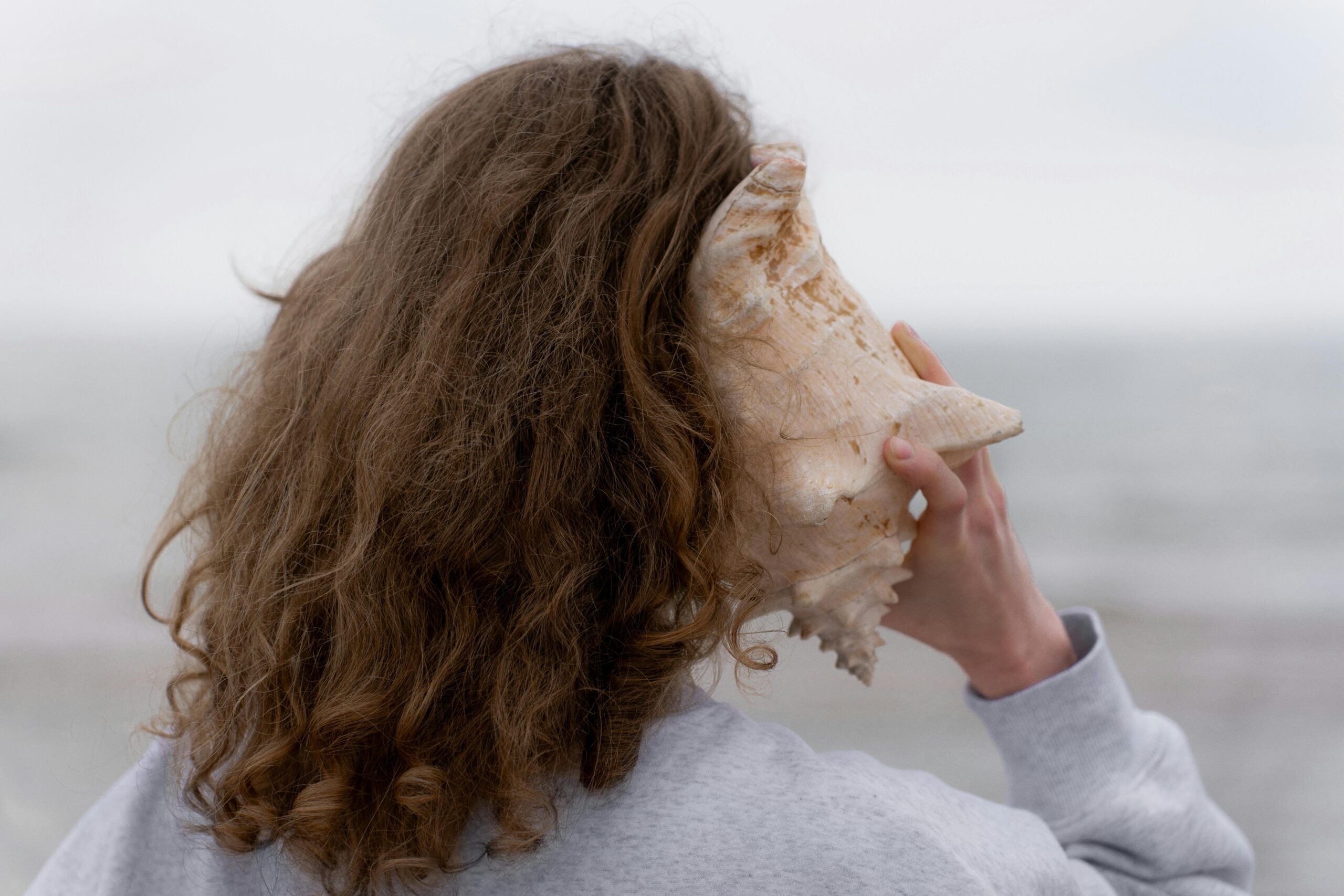 Eine Person mit lockigem Haar hält eine Muschel ans Ohr, während sie am Strand steht und auf das Meer blickt.