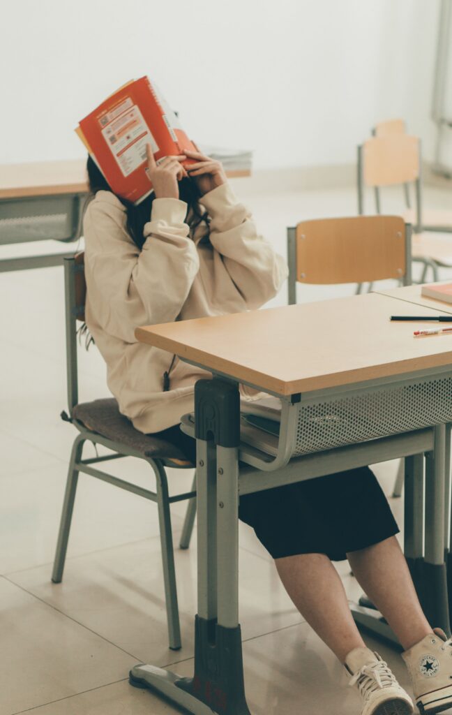 Junge Person sitzt an einem Tisch und hält ein Buch vor dem Gesicht, um sich zu verstecken.