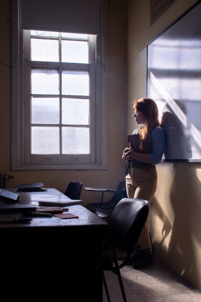 Eine Person mit langen roten Haaren steht nachdenklich neben einem Fenster in einem Klassenzimmer, umgeben von Schulmöbeln.